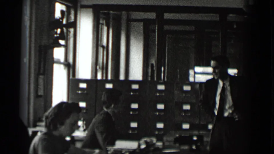 In the 1939 New York office scene, adults work and converse near filing cabinets, desks, tall windows, and an interior door.