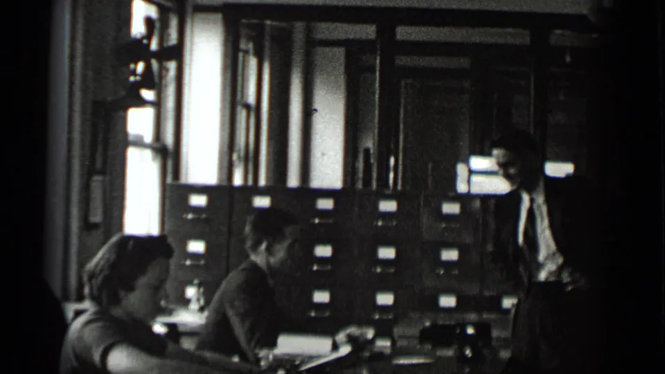 Black-and-white 1939 New York office footage shows seated adults at desks with a suited man standing by filing cabinets.