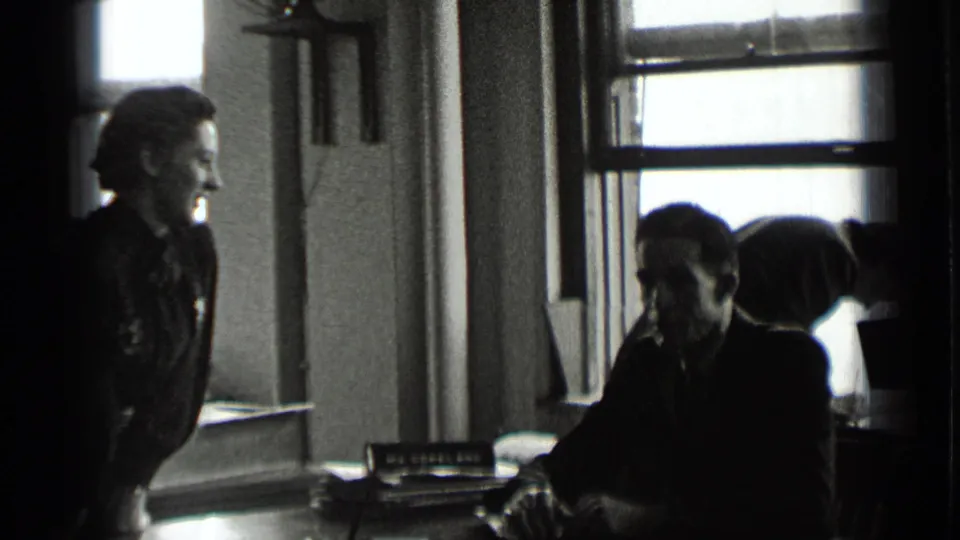 In the New York office interior, a woman stands near a window while seated adults appear to converse at a desk.