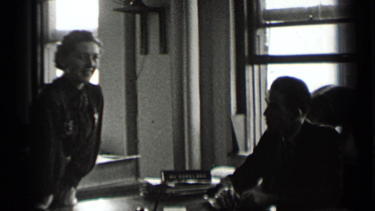 Woman standing near a desk while a seated man works in a window-lit office.