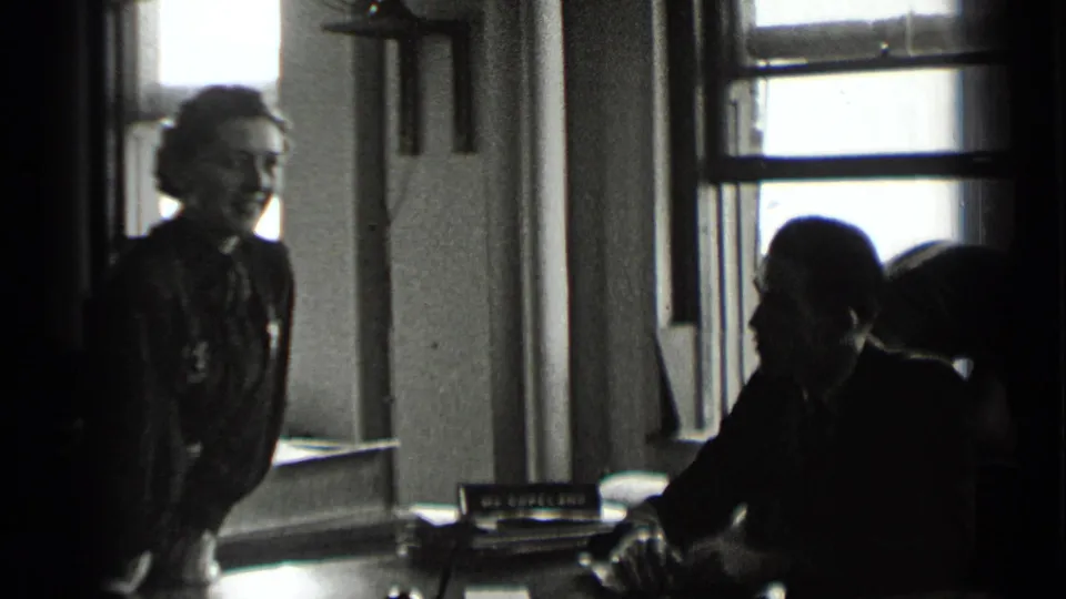 In a 1939 New York office scene, a woman in dark clothing stands by a desk as a seated suited man faces paperwork near tall windows.