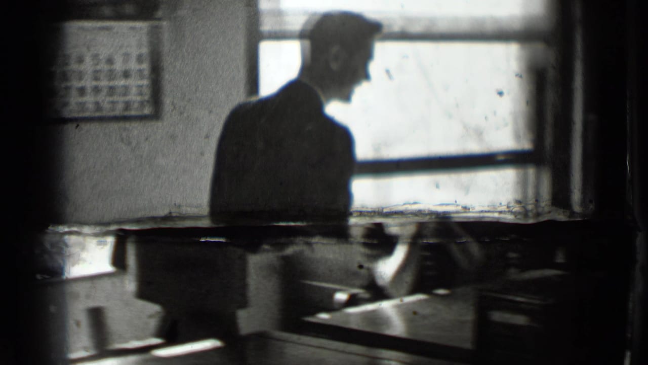A suited man stands in profile beside a bright window in the New York office interior, with desks and wall fixtures partly visible.