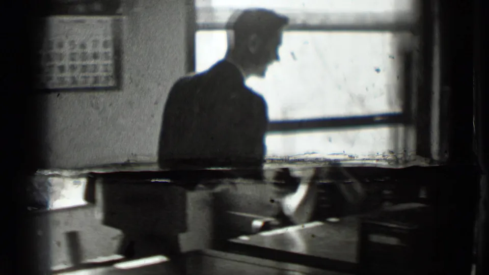 A suited man stands in profile beside a bright window in the New York office interior, with desks and wall fixtures partly visible.