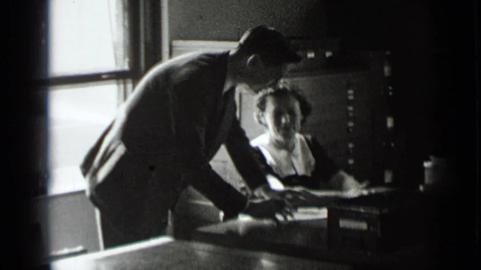 A man in a suit leans over a desk while a seated woman looks up, with filing cabinets and office papers visible behind them.