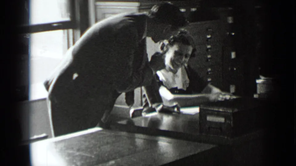A suited man bends over documents at a desk while a seated woman writes or reviews papers in the office interior.