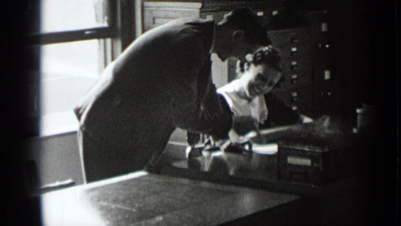 Man in a dark suit leans over a seated woman at an office desk.