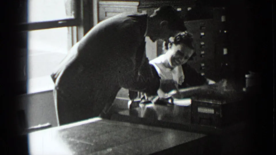 In a 1939 New York office scene, a suited man leans over a seated woman holding papers at a desk near a window.