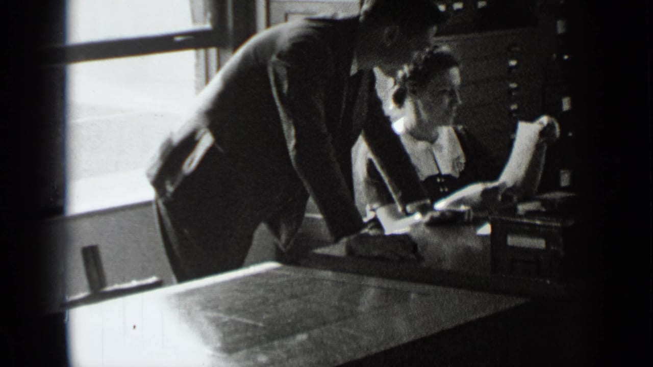 Suited man bends over a woman at a desk while she looks up with papers in hand.