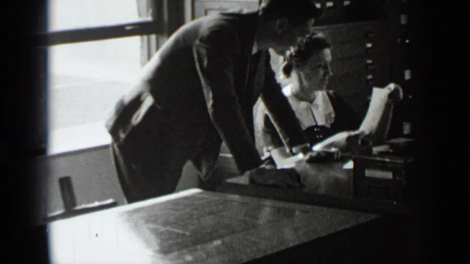 The man bends over the desk as the woman looks up from papers in the 1939 New York office interior.