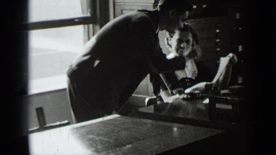 A woman seated at an office desk reacts toward the camera or man as he leans in beside her in the New York office scene.