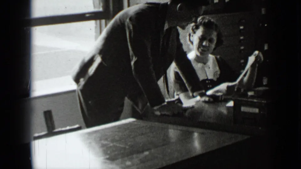 At the end of the clip, the suited man remains bent over the desk as the seated woman holds papers near office drawers.