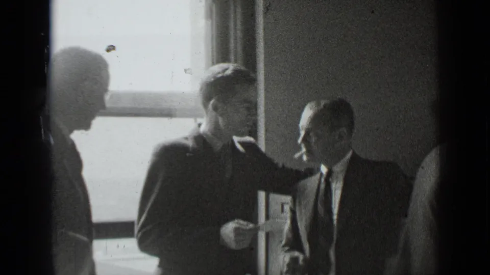 1939 New York home-movie frame showing men in suits conversing beside a bright window in a dim room.