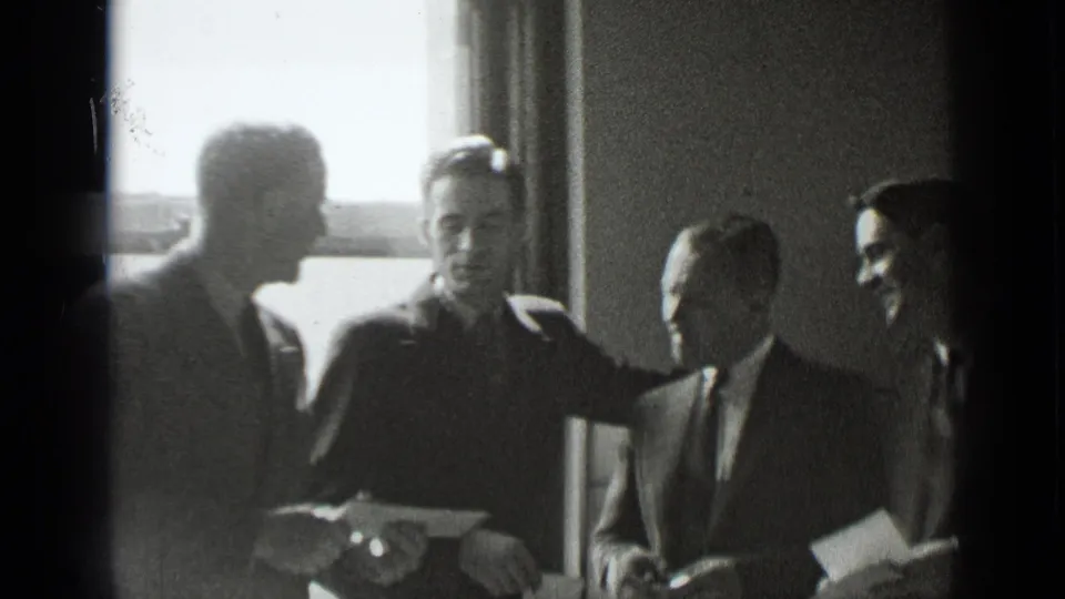 1939 New York home-movie frame showing four suited men gathered indoors, with papers visible and window light behind them.