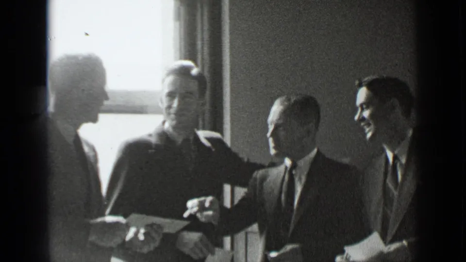1939 New York frame showing four suited men in conversation near a bright window, with one figure gesturing toward another.