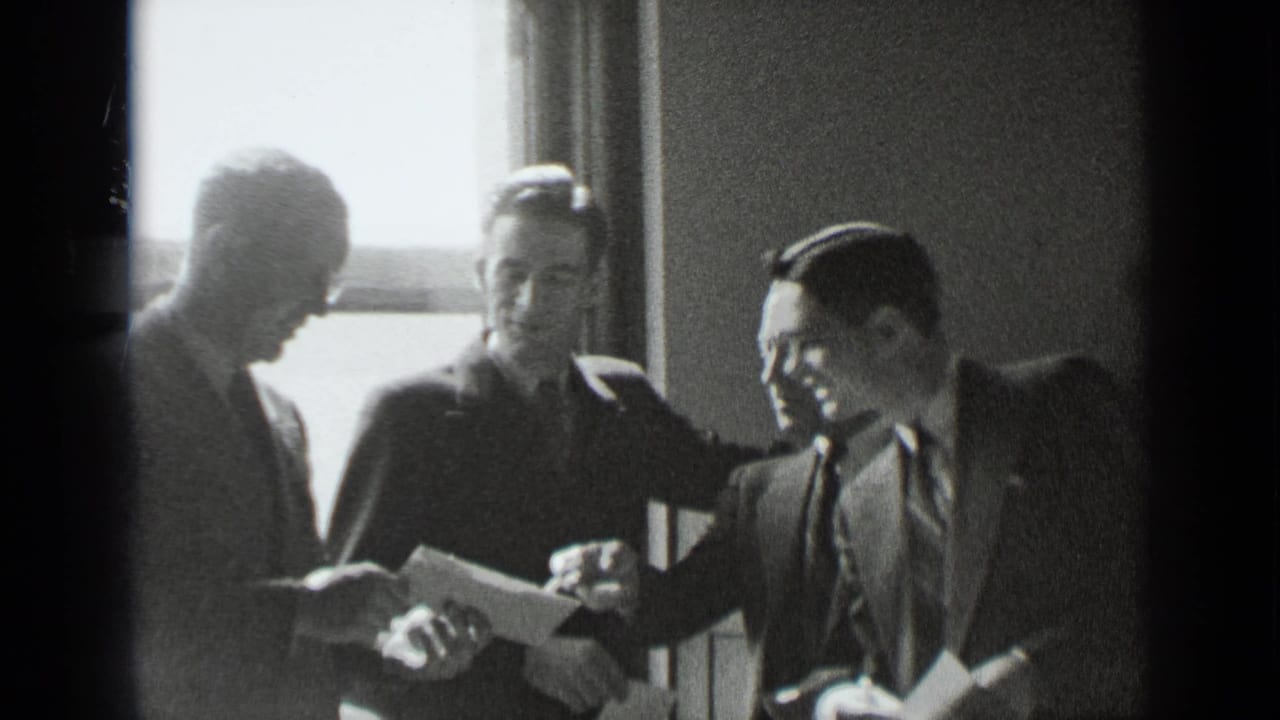 Four suited men stand or sit near a window while one holds papers.