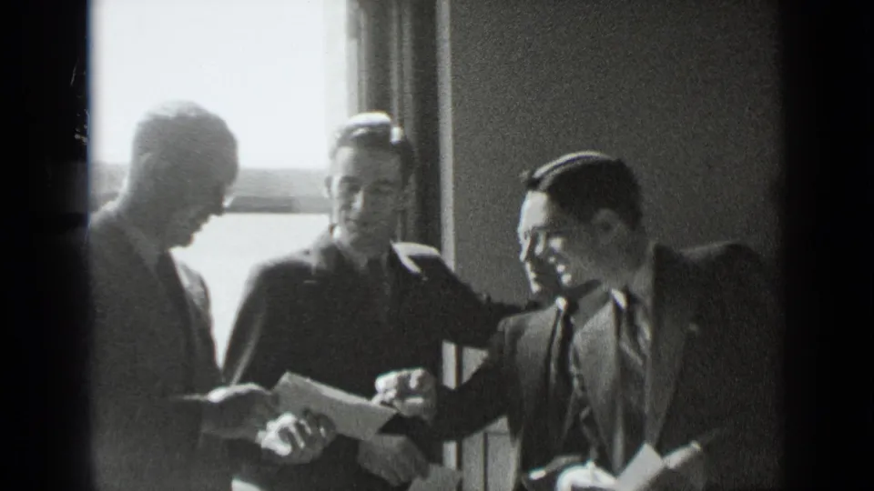 In 1939 New York home-movie footage, four suited men gather indoors near a bright window as papers are reviewed.