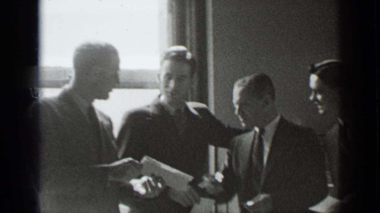 Four men in suits face each other as a paper is held open.