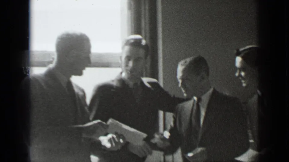 Four suited men in a New York indoor setting appear to confer over an open document near a bright window.