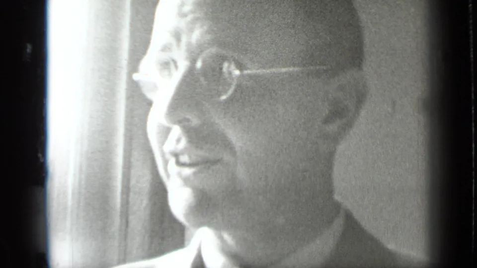 Close-up of a smiling man in round eyeglasses and a suit, backlit beside a window in the 1939 New York interior scene.