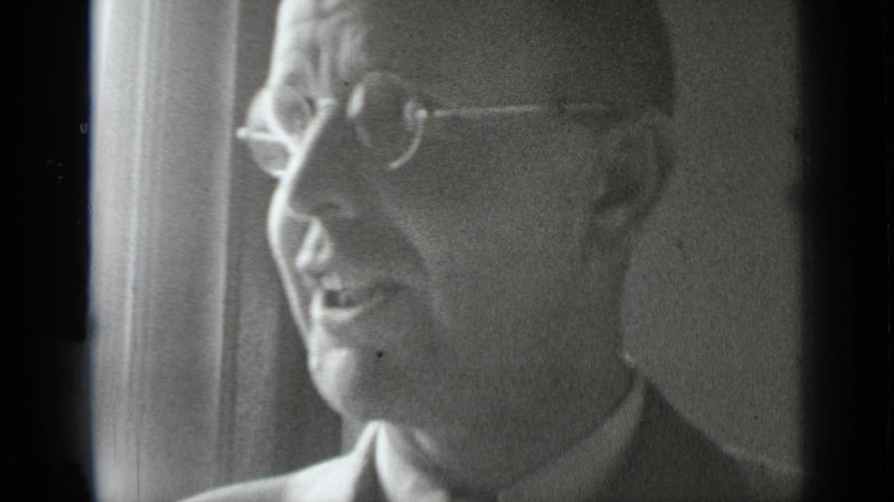 Older man in round glasses speaking in profile near a window.