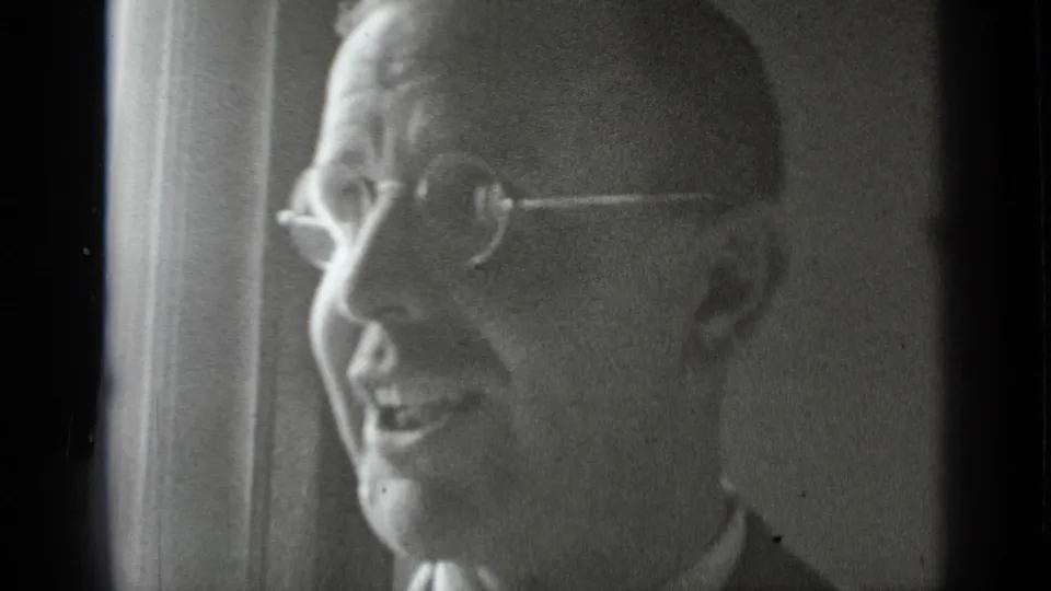 The man remains in profile, smiling or talking, with round glasses and soft window light in the black-and-white frame.