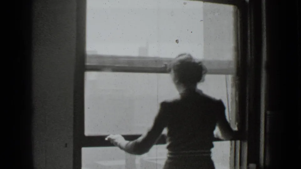 A woman in a dark dress stands with her back to the camera by a large bright window in the supplied New York home-movie setting.