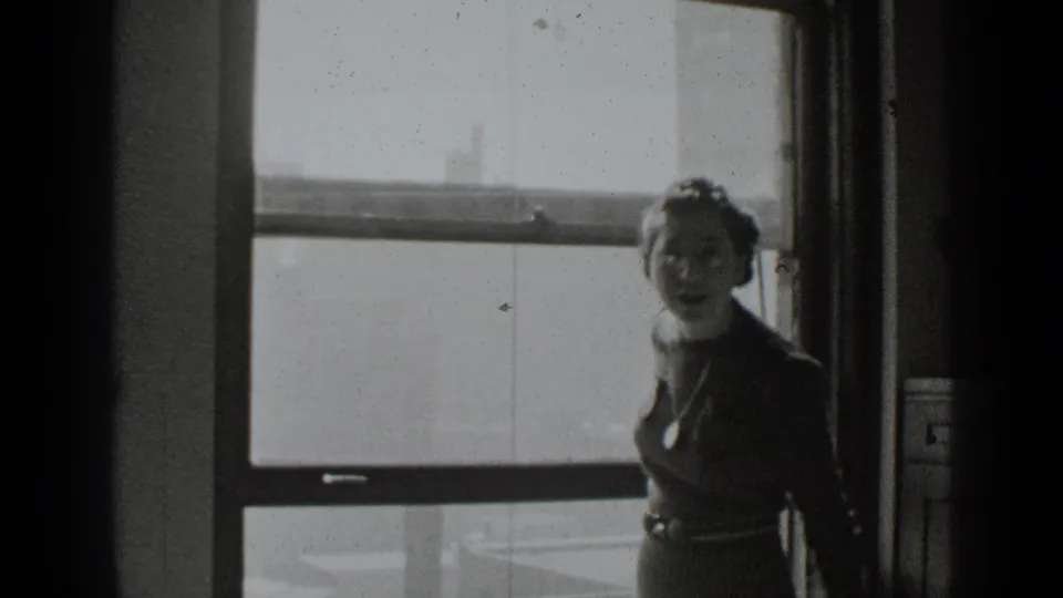 A woman in a dark dress turns toward the camera near a large window, her face visible in grainy black-and-white 8mm footage.