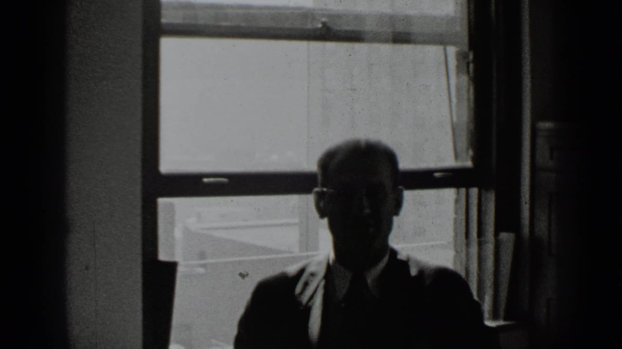 Backlit man in glasses seated in front of a large window.
