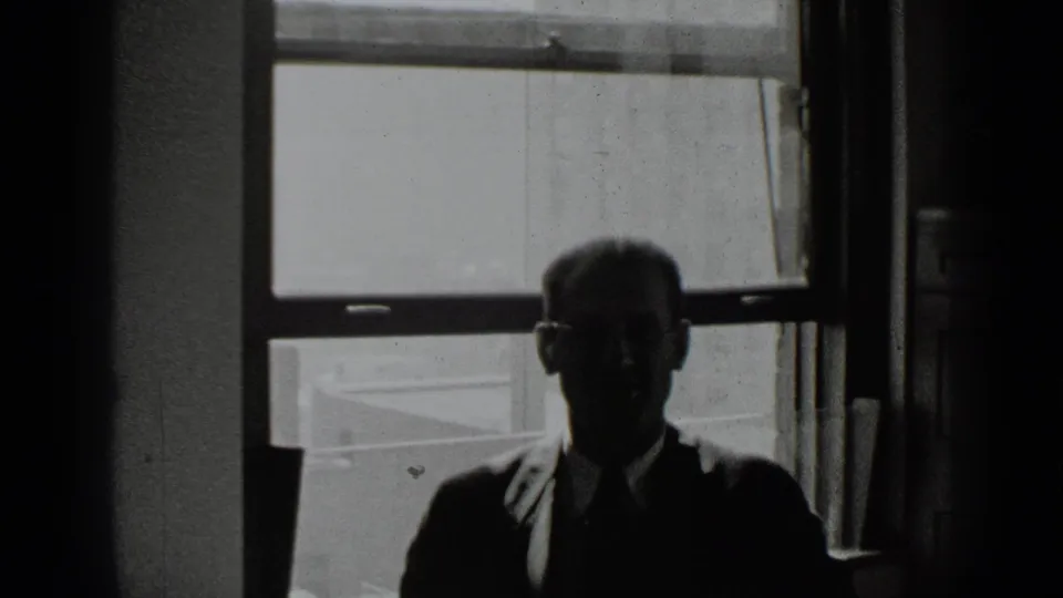 In 1939 New York home-movie footage, a backlit man in glasses sits before a bright window in a dim interior.