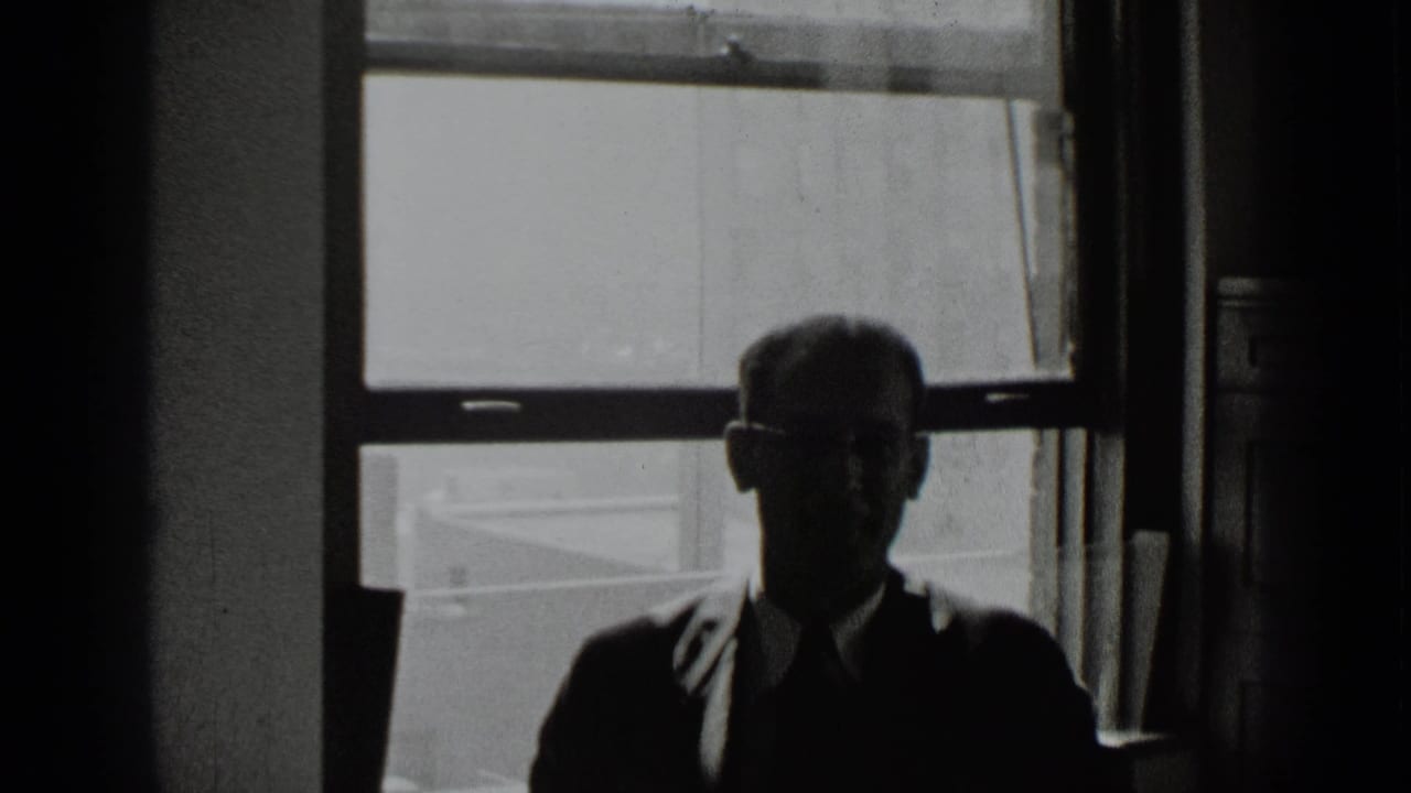 The man remains seated in shadow, with glasses and formal clothing visible against the bright window behind him.