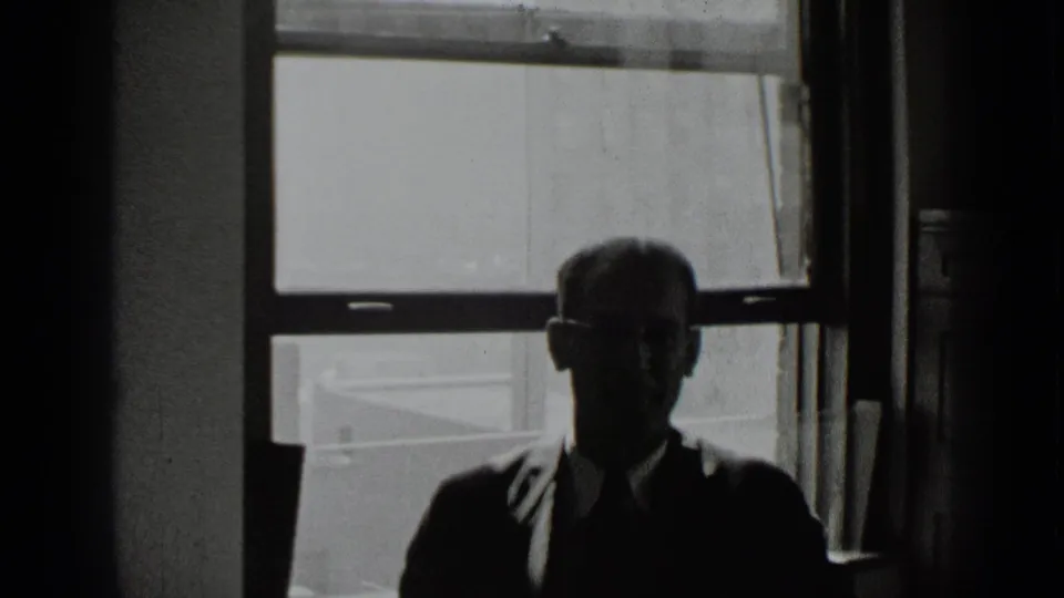 The man remains seated in shadow, with glasses and formal clothing visible against the bright window behind him.