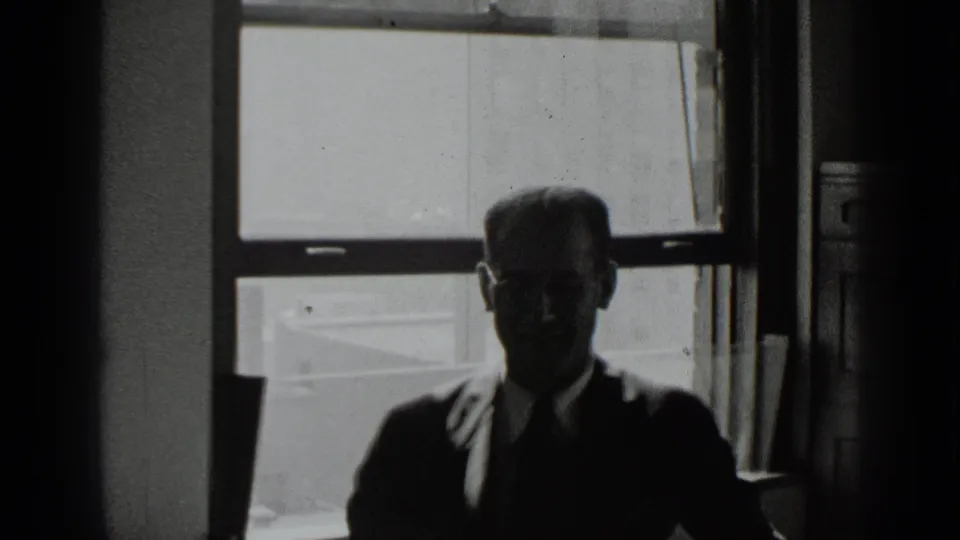 A seated man in glasses and a tie is partly illuminated while framed by the window and dark interior edges.