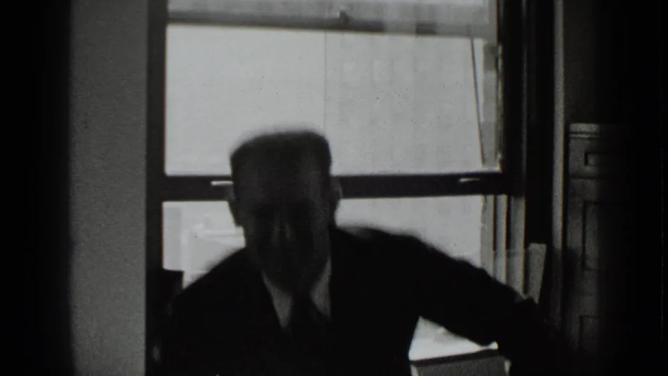 In a 1939 New York office scene, a man in a dark suit is silhouetted as he rises or leans near large windows.