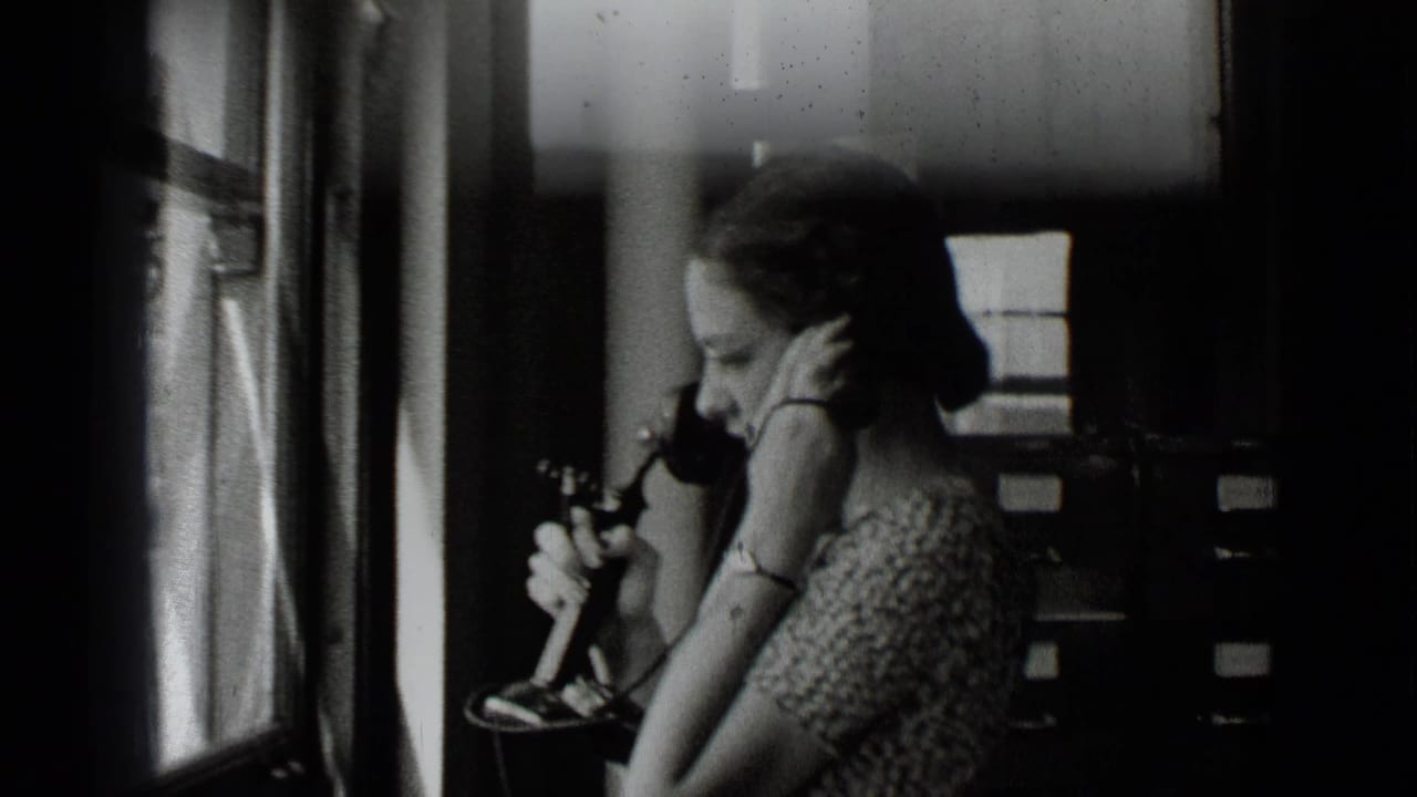 A woman in a patterned dress uses a desk telephone in a New York office, with windows and filing cabinets behind her.