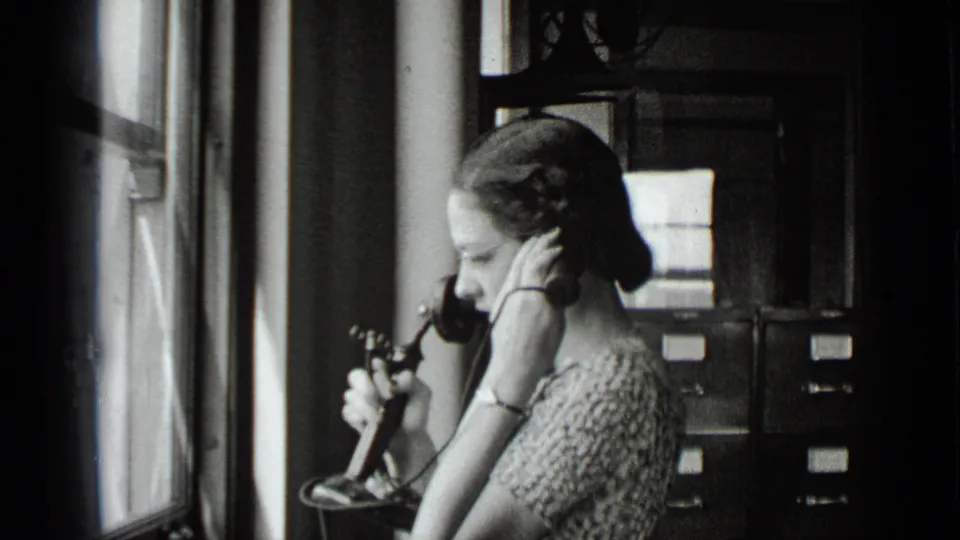 The woman continues the call beside a bright window, holding the receiver close with filing cabinets in the background.