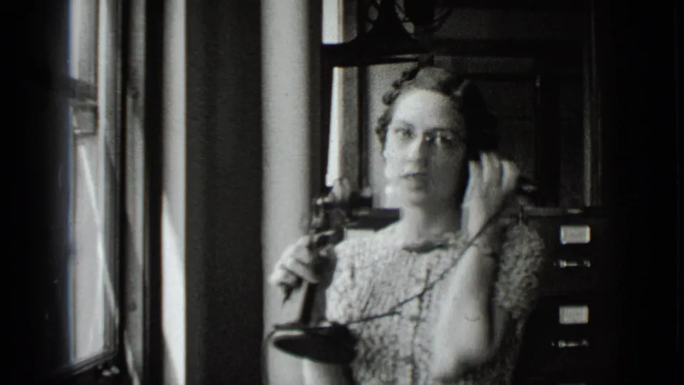 The woman turns toward the camera with the telephone receiver in hand, framed by office windows and filing cabinets.