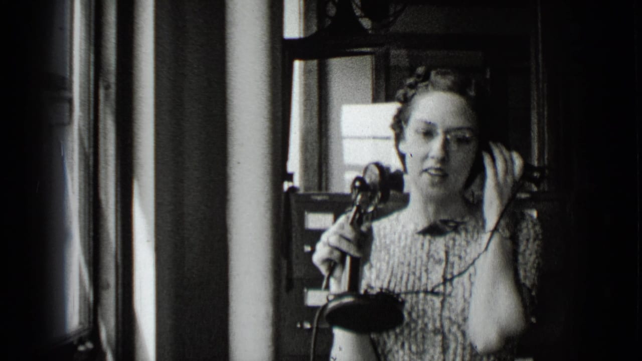 In 1939 New York footage, a woman in a patterned dress holds a vintage telephone receiver while standing or sitting in a dim office-like interior.