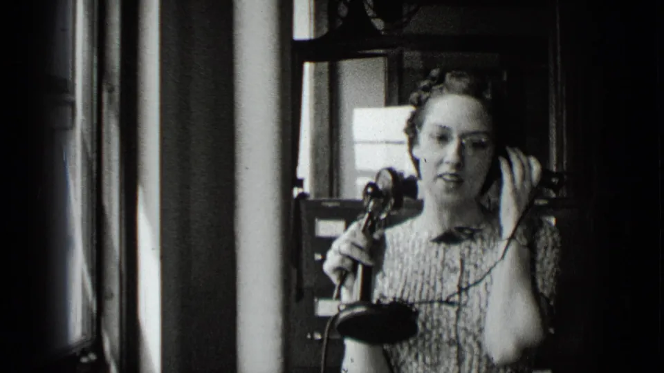 In 1939 New York footage, a woman in a patterned dress holds a vintage telephone receiver while standing or sitting in a dim office-like interior.