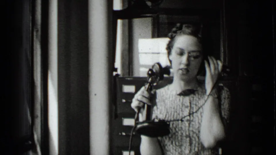 The woman appears to listen during a telephone call, holding the receiver and cord in a softly lit 1939 New York interior.