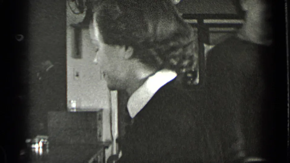 A woman is seen in side profile in the same black-and-white interior, with furniture and wall details partly visible behind her.