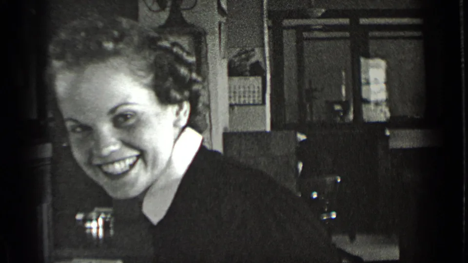 A smiling woman looks toward the camera in the 1939 New York home-movie footage, with paneled walls, furniture, and a doorway behind her.