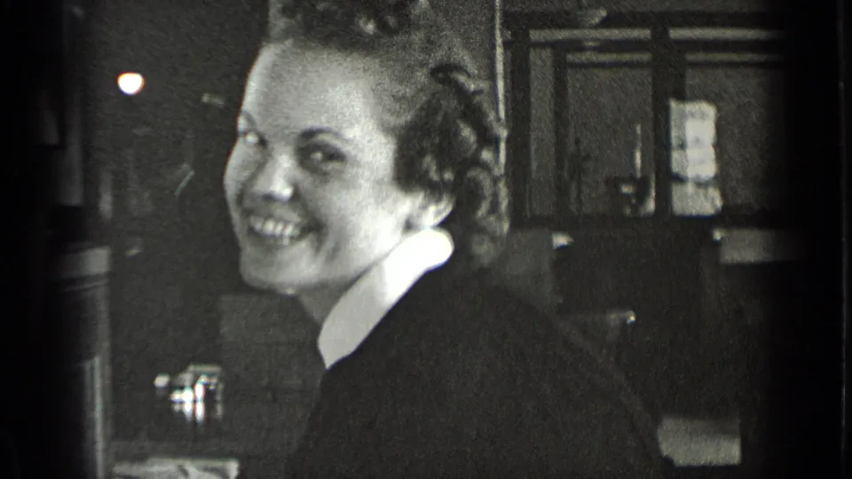 In supported 1939 New York footage, a woman in a dark dress with a white collar smiles near a workplace sink.