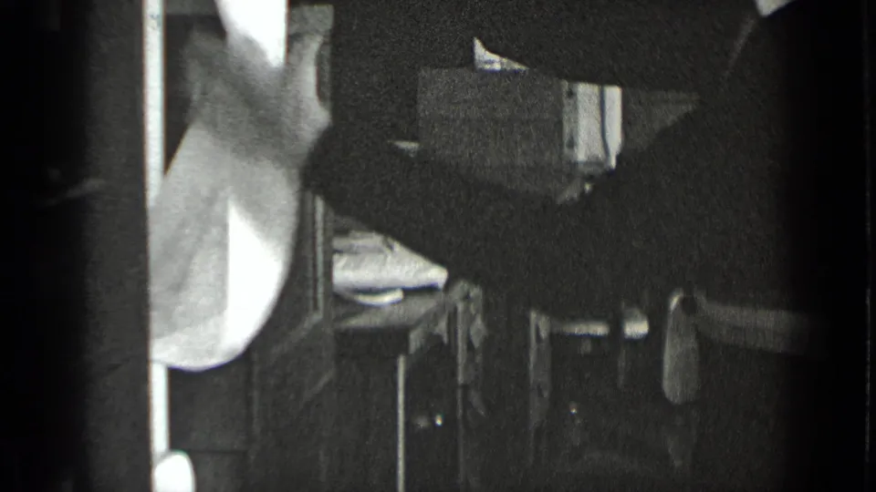 The frame shows the woman from the side at a sink, with a bright towel and dark workroom surfaces around her.