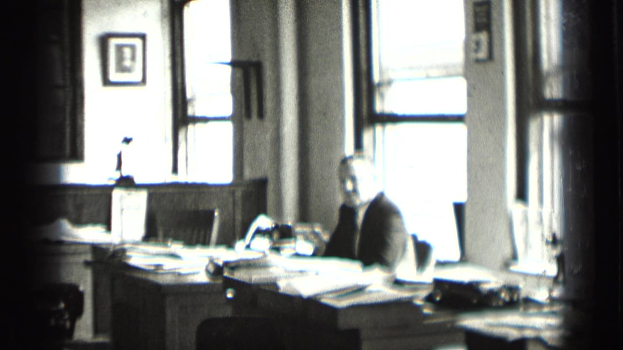 In a supported 1939 New York office scene, a suited man sits at a paper-covered desk with large windows and a framed portrait behind him.
