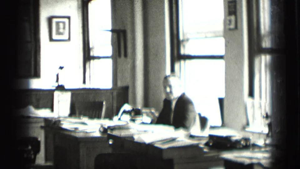 In a supported 1939 New York office scene, a suited man sits at a paper-covered desk with large windows and a framed portrait behind him.