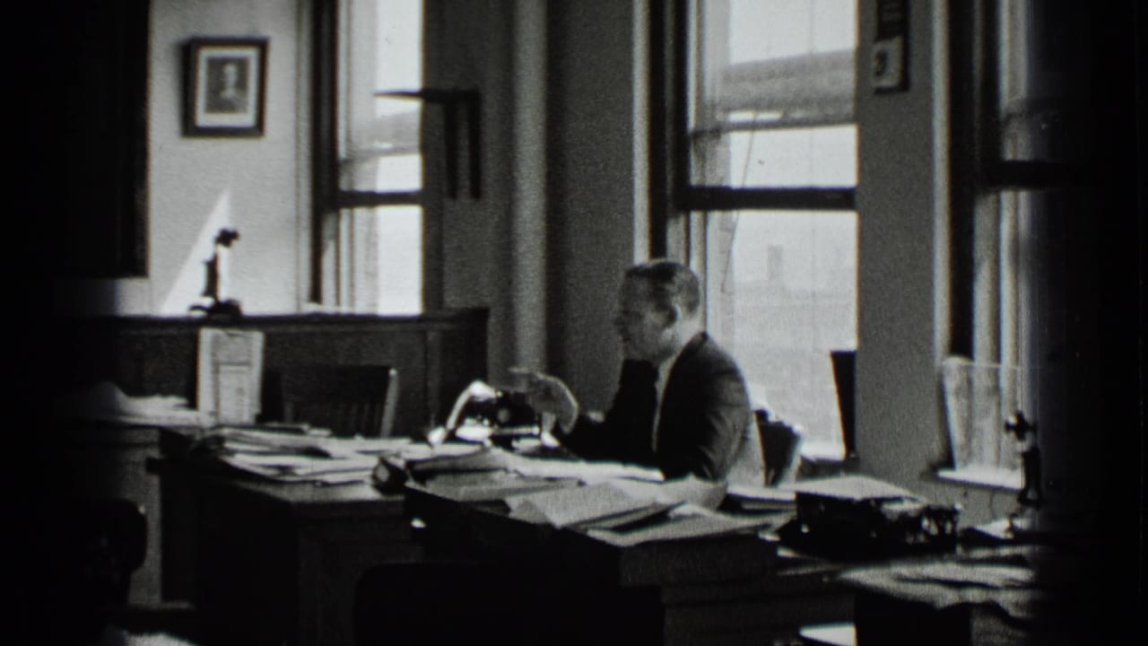 A suited man sits in profile at a crowded office desk in the supported New York 1939 footage, with papers, a manual typewriter, and tall windows visible.