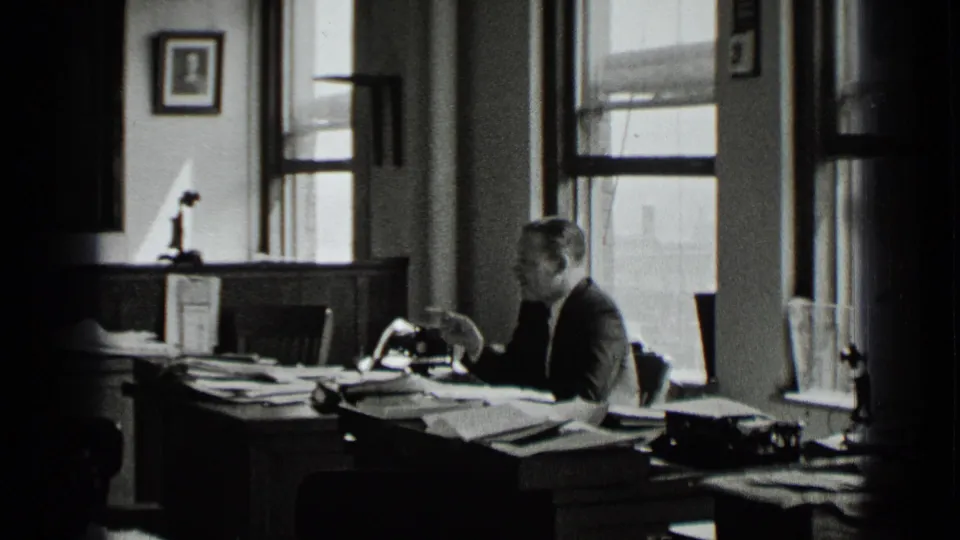 A suited man sits in profile at a crowded office desk in the supported New York 1939 footage, with papers, a manual typewriter, and tall windows visible.