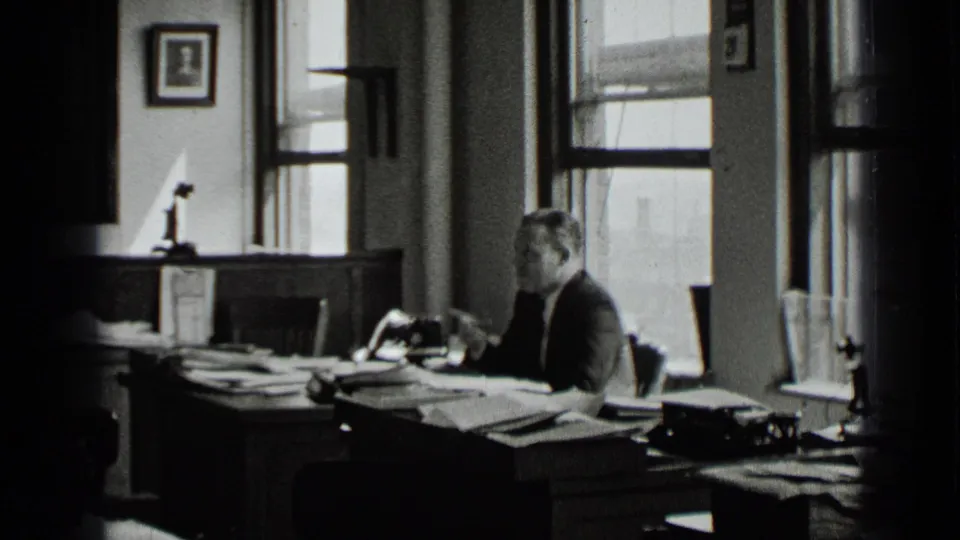 The frame shows the man seated beside a document-covered desk in the supported 1939 New York office interior, with windows, wall art, and desk equipment in view.