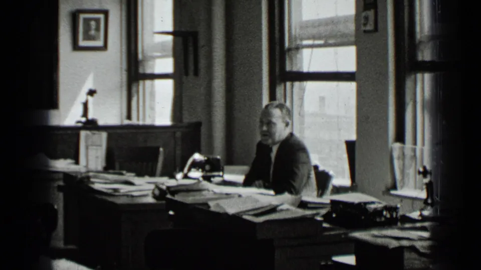 A suited man turns more toward the camera while seated at a cluttered desk in the supported 1939 New York office scene, with papers, windows, and a typewriter visible.
