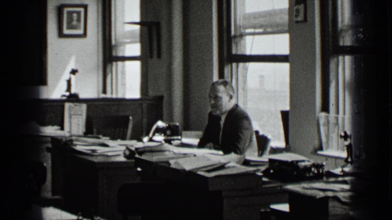 Man in a suit sits at a cluttered office desk near windows and papers.